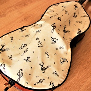 May include: A beige cello case with a black musical note pattern. The case is made of a soft, fabric-like material and has a zipper closure.