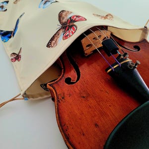 May include: A violin is partially enclosed by a cream-coloured case decorated with colourful butterfly illustrations. The violin's wooden body is a rich reddish-brown, with strings and a bridge visible. The case has a drawstring closure.