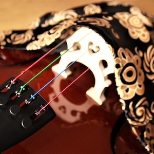 May include: Close-up of a cello with four strings, a black tailpiece, and a white bridge. The cello is covered with a black and gold patterned fabric.