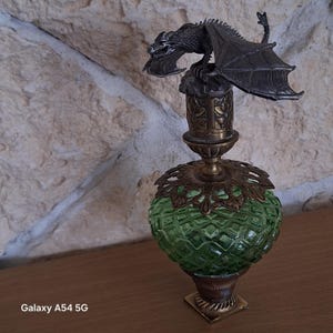 Peut inclure: Un flacon de parfum en verre vert avec une texture à motif losange, doté d'une base en laiton et d'un col en métal orné. Le bouchon est surmonté d'une figurine de dragon argenté détaillée aux ailes déployées, posée sur un petit rocher.
