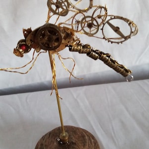 May include: A gold metal dragonfly sculpture with intricate clockwork details, including gears and wheels, on a wooden base.