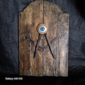 Puede incluir: Una placa de pared rectangular con un acabado texturizado y desgastado en bronce y negro. Centrado en la superficie hay un ojo azul realista dentro de un engranaje metálico, colocado sobre un par de compases de dibujo de metal negro cruzados.