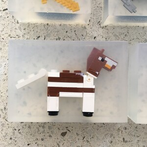 May include: A white and brown Lego horse figurine with a brown head and tail. The horse is standing on a clear plastic base.
