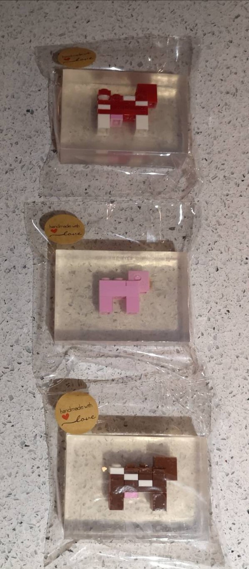 May include: Three clear rectangular bars of soap with a handmade with love label. Each bar features a different animal made of colorful Lego blocks. The first bar features a red and white cow, the second bar features a pink pig, and the third bar features a brown and white cow.