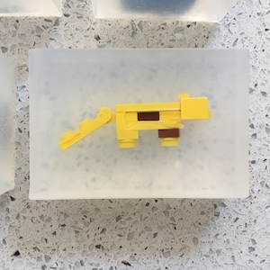 May include: A yellow plastic toy animal with a brown stripe on its back. The toy is made of Lego bricks and is sitting on a clear surface.