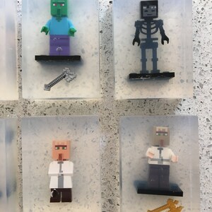May include: Four clear plastic boxes each containing a different Minecraft character figurine. The figurines are a green zombie, a black skeleton, a villager with a white shirt, and a villager with a brown shirt. Each box also contains a small accessory, such as an axe or a sword.