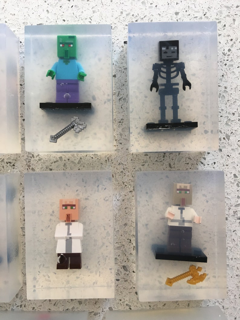 May include: Four clear plastic boxes each containing a different Minecraft character figurine. The figurines are a green zombie, a black skeleton, a villager with a white shirt, and a villager with a brown shirt. Each box also contains a small accessory, such as an axe or a sword.