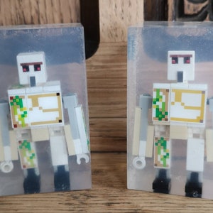 May include: Two clear resin blocks, each containing a white Lego Minecraft figure with red eyes and a gold and green patterned chest plate. The figures are posed standing with their arms outstretched.