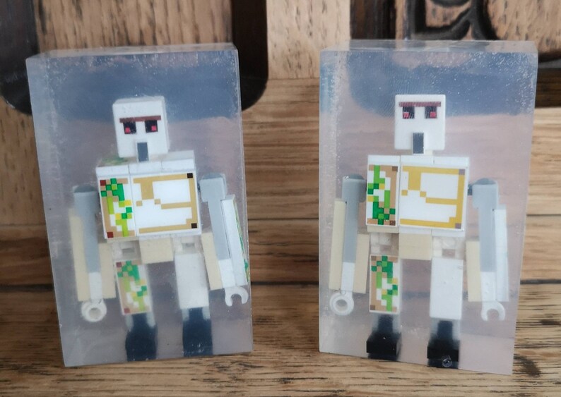 May include: Two clear resin blocks, each containing a white Lego Minecraft figure with red eyes and a gold and green patterned chest plate. The figures are posed standing with their arms outstretched.