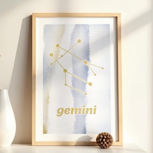 Op de afbeelding: Een ingelijste verticale wandkunstprint met een aquarelachtergrond met verticale strepen in tinten blauw, grijs en beige. De print toont een goudkleurig sterrenbeeld-diagram van Tweelingen en het woord gemini in gouden letters.