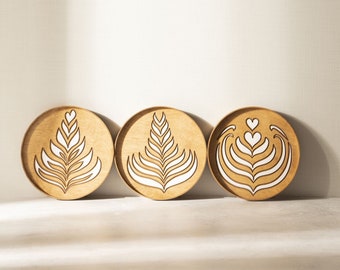 May include: Three round wooden trays with white latte art designs. The trays are arranged in a row on a light brown surface.