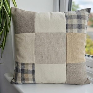 May include: Square cushion with a patchwork design. The cushion features a variety of neutral-toned fabrics, including beige, cream, and grey, arranged in a grid pattern. Some squares have a herringbone or plaid texture.