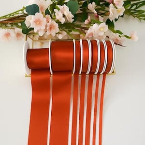 Könnte beinhalten: Mehrere Rollen rostfarbenes Satinband mit weißen Kanten, angeordnet in einem goldfarbenen Metallhalter. Das Band ist abgewickelt, wobei mehrere Stränge herunterhängen. Weiche rosa und weiße Blumen sind im Hintergrund.