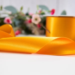 K&ouml;nnte beinhalten: Eine Spule mit leuchtend gelbem Satinband, wobei ein St&uuml;ck Band abgewickelt ist und auf einer wei&szlig;en Oberfl&auml;che liegt.