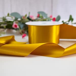 K&ouml;nnte beinhalten: Eine Spule mit goldenem Satinband, auf der ein St&uuml;ck Band aufgewickelt ist. Das Band ist glatt und gl&auml;nzend.