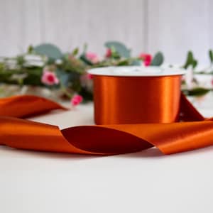 K&ouml;nnte beinhalten: Eine Spule mit orangefarbenem Satinband, auf der ein St&uuml;ck Band auf einer wei&szlig;en Oberfl&auml;che liegt.
