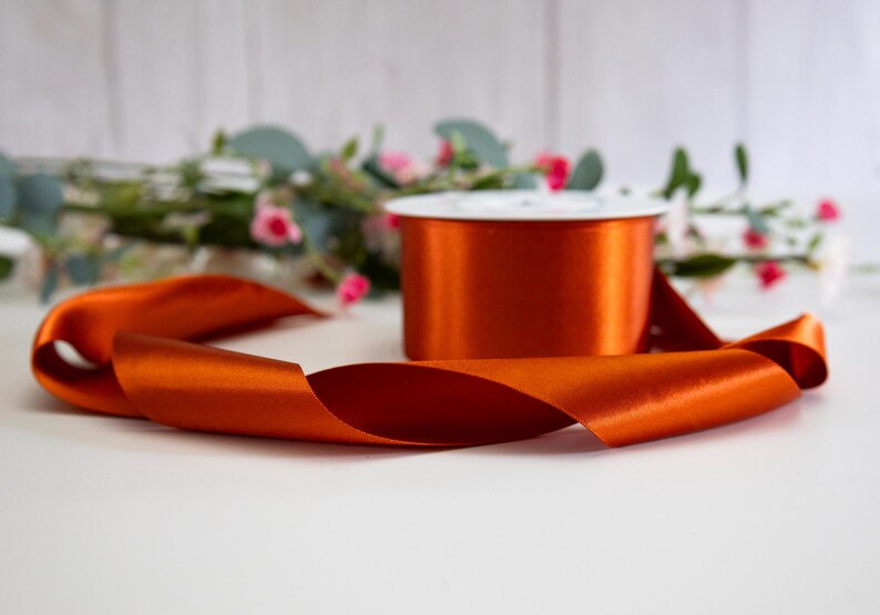 K&ouml;nnte beinhalten: Eine Spule mit orangefarbenem Satinband, auf der ein St&uuml;ck Band auf einer wei&szlig;en Oberfl&auml;che liegt.