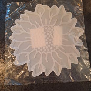 Peut inclure: Un moule en silicone translucide blanc cassé en forme de tournesol avec des contours de pétales détaillés et un centre texturé. Le moule est contenu dans un sac en plastique transparent, présentant une surface plate et flexible.