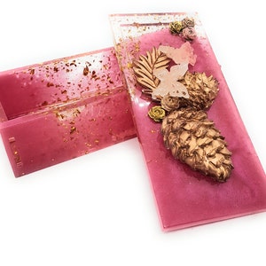 Può includere: Una scatola rettangolare rosa con una finitura glitter dorata. Il coperchio della scatola è aperto e presenta una pigna dorata, foglie dorate e farfalle rosa. La scatola è perfetta per riporre piccoli oggetti.
