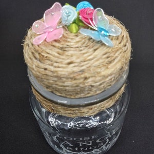 Könnte beinhalten: Ein klares Glasgefäß mit einem Deckel aus Jute und einer dekorativen Schnur. Der Deckel ist mit drei Stoffschmetterlingen in Rosa, Blau und Grün verziert. Das Gefäß trägt die Worte "Loose Change" und "Ring", die in das Glas geätzt sind.