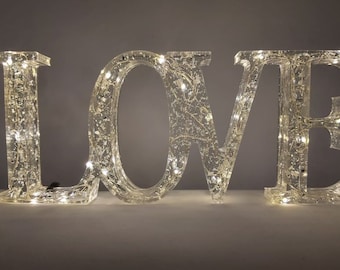 May include: A clear plastic sign spelling out the word "LOVE" with white lights embedded in the plastic.