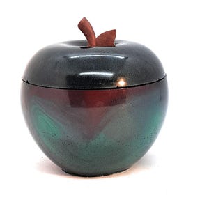 May include: A ceramic apple-shaped container with a lid. The container is black, red, and green with a swirling pattern. The lid is a dark brown and has a leaf-shaped handle.