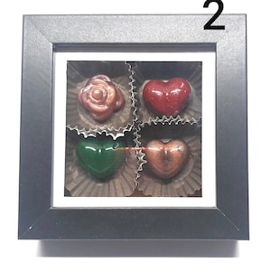 Peut inclure: Une boîte noire encadrée avec une fenêtre transparente montrant quatre bonbons au chocolat. Les bonbons sont en forme de rose et de cœurs. Les bonbons sont de différentes couleurs, notamment rouge, vert et marron.