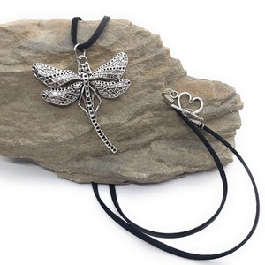 Può includere: Una collana con un ciondolo a forma di libellula argentata su un cordoncino in pelle nera. La collana ha una chiusura a forma di cuore.