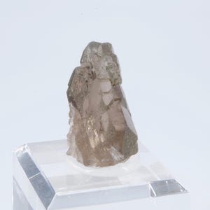 May include: A smoky quartz crystal with a natural, rough texture. The crystal is a light brown color and has a pointed shape. It is displayed on a clear acrylic stand.