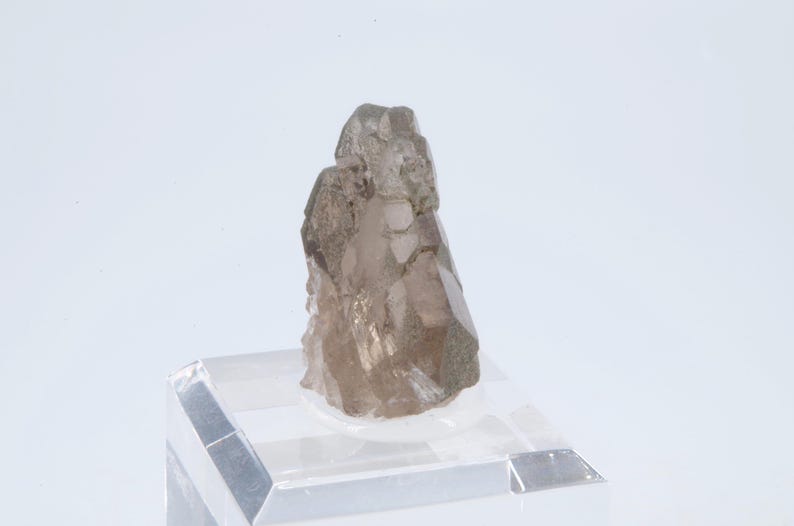 May include: A smoky quartz crystal with a natural, rough texture. The crystal is a light brown color and has a pointed shape. It is displayed on a clear acrylic stand.