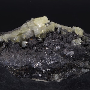 Puede incluir: Un grupo de cristales de color amarillo pálido sobre una matriz gris oscuro. Los cristales son translúcidos y tienen un brillo vítreo. La matriz es rugosa y texturizada.