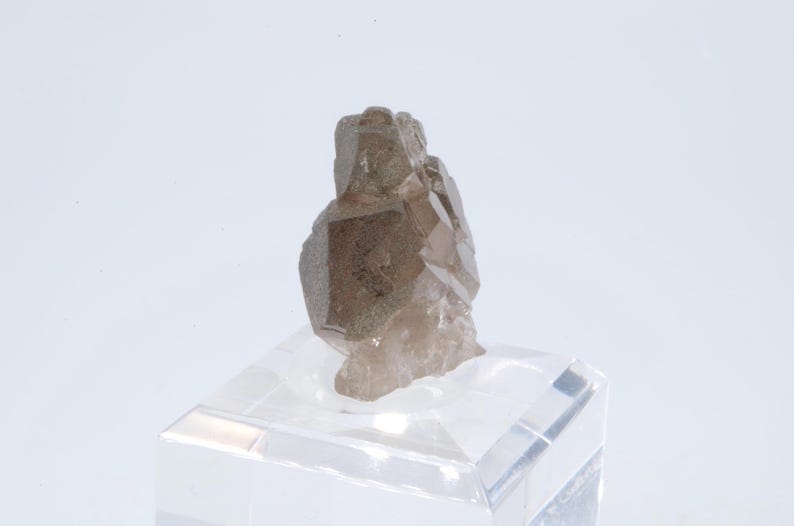 May include: A smoky quartz crystal cluster with a natural, rough texture. The crystal is a light brown color and has a translucent appearance. The crystal is displayed on a clear acrylic base.
