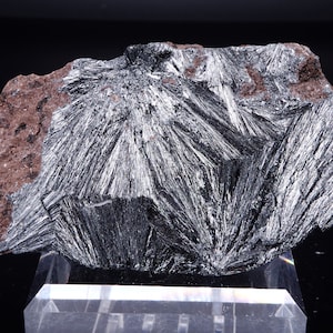 May include: A large, gray, mineral specimen with a rough, textured surface. The mineral is composed of many thin, needle-like crystals that are arranged in a radial pattern. The specimen is displayed on a clear acrylic base.
