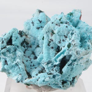 Peut inclure: Spécimen minéral saisissant, présentant une formation bleu clair texturée et vibrante, avec des inclusions vert foncé et noires. La structure cristalline a un aspect poreux, créant un attrait visuel unique. Le minéral est exposé sur un support transparent.