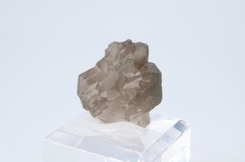 May include: A cluster of smoky quartz crystals with a light pink hue. The crystals are translucent and have a rough, natural texture. The cluster is displayed on a clear acrylic base.