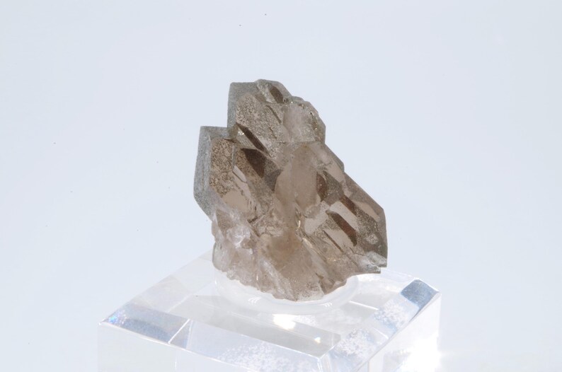 May include: A smoky quartz crystal cluster with a natural, rough texture. The crystal is a light brown color and has a pointed shape. The crystal is displayed on a clear acrylic base.