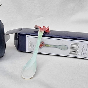 Puede incluir: Una cuchara de porcelana blanca con un diseño de flor en el mango. La cuchara está en su embalaje original, que es una caja azul con texto blanco.