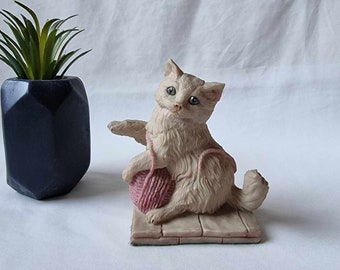 May include: White ceramic figurine of a cat sitting on a wooden platform, holding a pink ball of yarn. The cat has blue eyes and a playful expression.