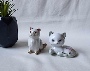 May include: Two white porcelain cat figurines. One cat is sitting upright with gold accents and the other is lying down with pink floral accents.