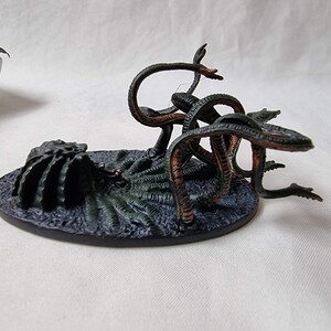 May include: A green and brown painted miniature of a multi-headed hydra monster with multiple tentacles. The hydra is on a black base with a textured surface.