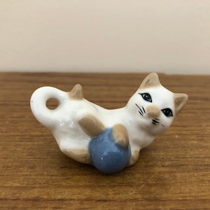 May include: A white ceramic cat figurine with brown markings, playing with a blue ball. The cat has black eyes and a cute expression.