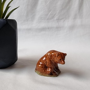 May include: A small brown ceramic bear figurine sitting on a white surface. The bear is looking down and has a brown nose and eyes.