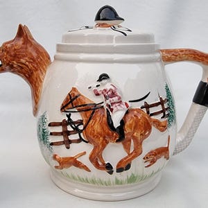 May include: A white ceramic teapot with a fox spout and a handle shaped like a fox's leg. The teapot is decorated with a scene of a rider on a brown horse jumping over a fence, with two smaller foxes in the background.