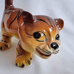 May include: A ceramic figurine of a tiger cub with brown and black stripes. The figurine has a playful expression with black eyes and a black nose.