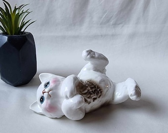 May include: White ceramic figurine of a cat lying on its back with its paws in the air. The cat has pink cheeks and blue eyes.