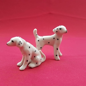 Op de afbeelding: Twee kleine keramische Dalmatiër-hondjes. Eén hondje staat en het andere zit. Beide hondjes zijn wit met zwarte vlekken.