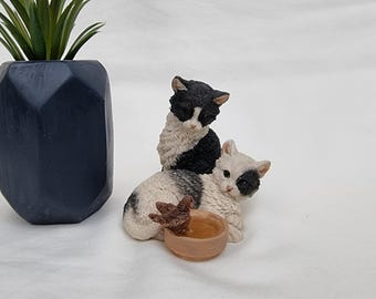 May include: A dark blue geometric planter holds a green plant. Two cat figurines are in the foreground. One cat is black and white, and the other is white with black spots. A small brown bowl is in front of the cats.