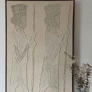 May include: A beige textured canvas painting with a brown wooden frame. The painting depicts two figures in profile, wearing elaborate headdresses and robes. The figures are holding staffs in their hands.