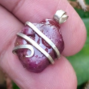 Peut inclure: Pendentif en rubis rouge foncé brut, enveloppé de fil d'argent. Le rubis, de forme irrégulière, présente une surface polie et brillante. Le fil d'argent est enroulé autour de la pierre, créant un bijou artisanal unique.