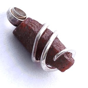 May include: A pendant featuring a rough, reddish-brown gemstone wrapped in a silver-colored wire. The pendant has a silver-colored bail at the top for a chain. The gemstone has a textured surface and irregular shape.